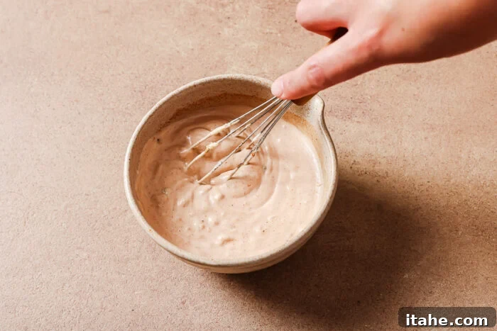 A hand whisking a creamy, light brown sauce or dressing in a beige ceramic bowl on a light brown surface.
