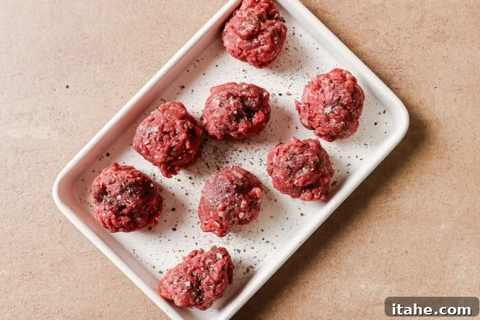 A white rectangular tray holds eight raw beef meatballs sprinkled with ground black pepper, set on a light brown surface.