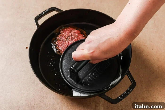 A hand presses a black cast iron burger press onto a raw beef patty in a black cast iron skillet on a tan surface.