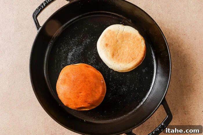 A cast iron skillet with two English muffins inside, one with a golden brown top and the other with a lighter, pale top, sitting on a beige surface.