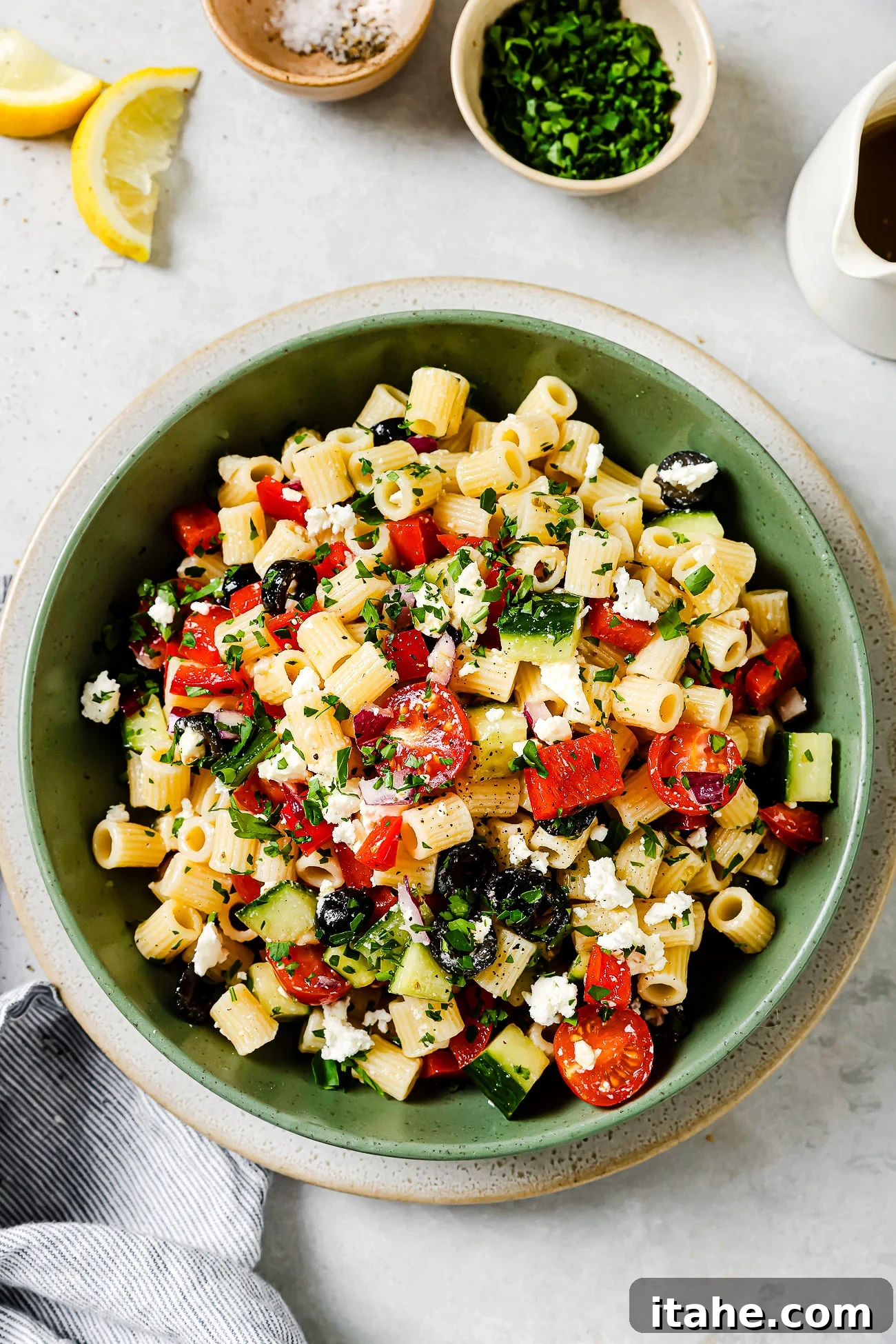 A green bowl filled with pasta salad containing cherry tomatoes, cucumbers, red peppers, black olives, feta cheese, and chopped herbs. Small bowls of salt and chopped herbs, lemon wedges, and a napkin are nearby.