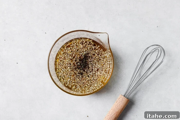 A glass measuring cup filled with oil and herbs sits on a light surface next to a metal whisk with a wooden handle.