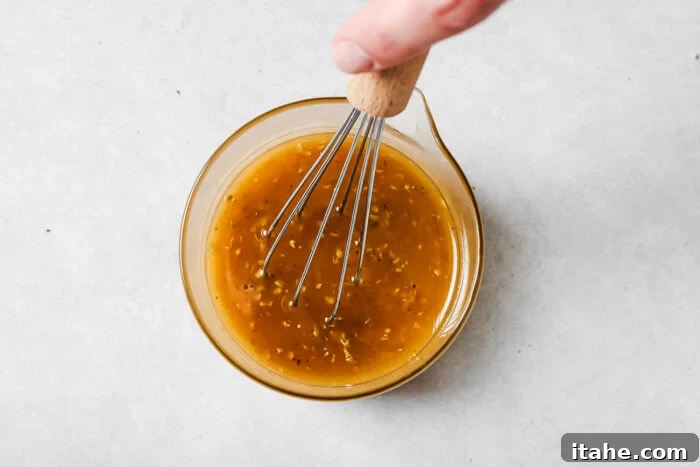A hand holding a whisk is mixing a yellow-brown liquid with herbs in a small glass bowl on a light gray surface.