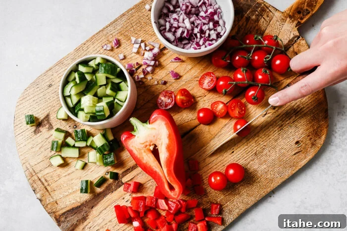 A hand slices cherry tomatoes on a wooden cutting board, surrounded by chopped cucumber, red bell pepper, diced red onion, and a bowl of whole and halved cherry tomatoes.