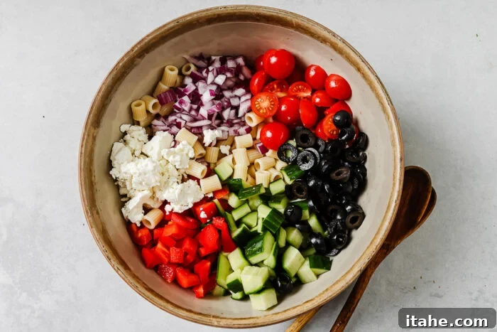 A large bowl containing cooked pasta, chopped red onions, cherry tomatoes, sliced black olives, cucumber, diced red bell pepper, and crumbled feta cheese, ready to be mixed into a salad. Wooden salad servers lie beside the bowl.