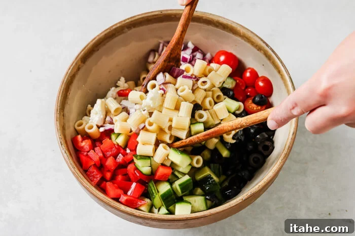 A person uses wooden utensils to mix a bowl filled with pasta, chopped red bell pepper, cucumber, cherry tomatoes, red onion, black olives, and feta cheese.