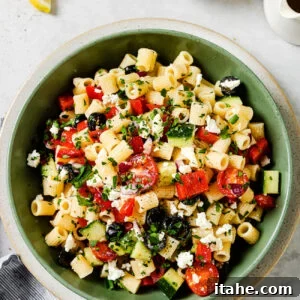 A green bowl filled with pasta salad containing cherry tomatoes, cucumbers, red peppers, black olives, feta cheese, and chopped herbs. Small bowls of salt and chopped herbs, lemon wedges, and a napkin are nearby.
