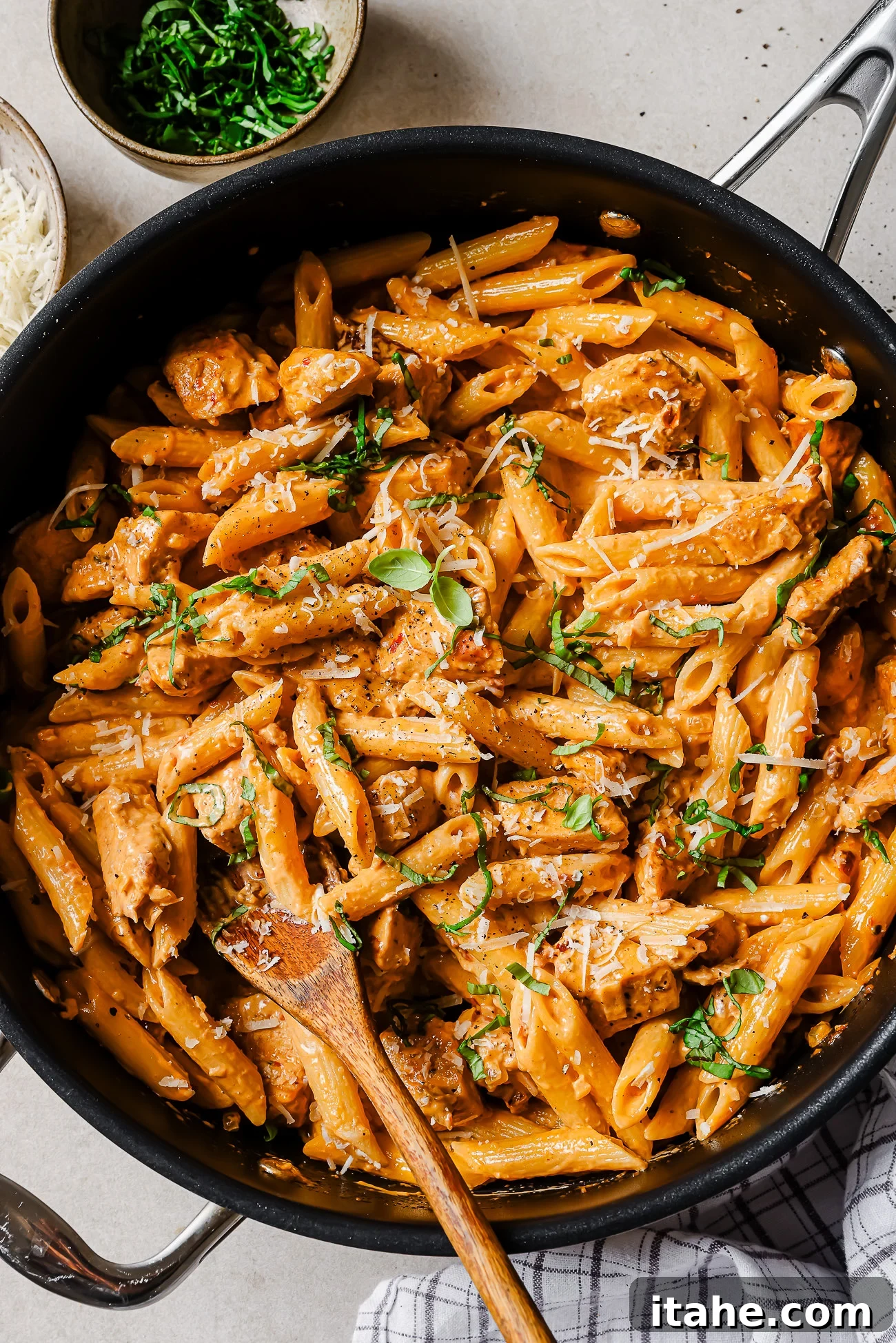 Creamy Marry Me Chicken Pasta Recipe 2 A large skillet filled with creamy penne pasta, pieces of chicken, fresh herbs, and grated cheese, with a wooden spoon for serving. Bowls of chopped herbs and grated cheese are nearby on the table.