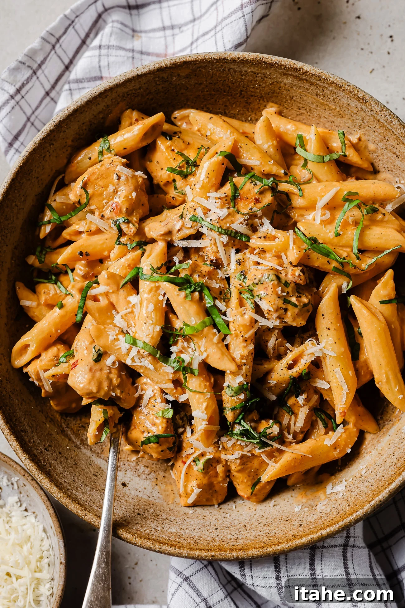 Creamy Marry Me Chicken Pasta Recipe 6 A bowl of creamy penne pasta with pieces of chicken, topped with grated cheese and chopped fresh herbs. A fork is placed in the bowl, and a checked cloth napkin is partially visible underneath.