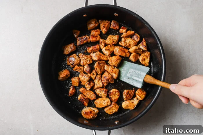 Creamy Marry Me Chicken Pasta Recipe 5 A hand holding a spatula stirs browned, seasoned chicken pieces in a black frying pan on a light-colored countertop.